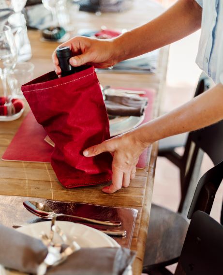 A person is shown removing a bottle of wine from a red washable paper wine holder, which sits atop a laid table.