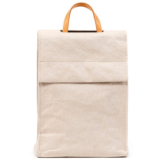 A tall rectangular square-edged washable paper backpack with a front flap and a tan handle is shown in a cream colour.