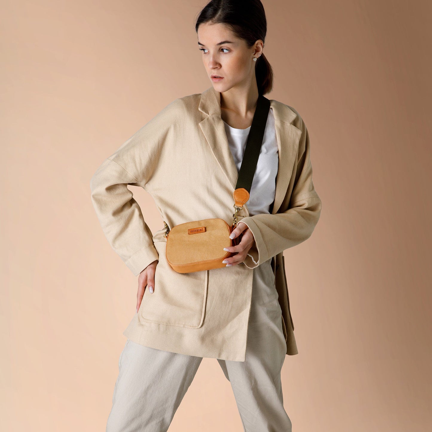 A woman stands in front of a peach-toned wall, wearing a beige blazer, white t-shirt and chinos. Across her body is a tan coloured washable paper bag with a dark cotton canvas strap.