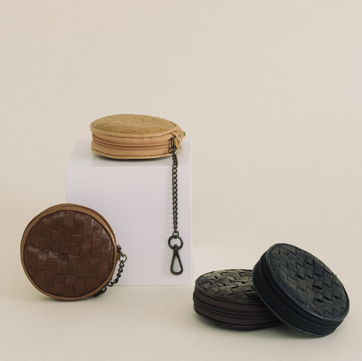 A tan washable paper woven coin purse is shown atop a small white plinth, with its antique brass attachment chain dangling down the edge. At left sits the same purse in mid brown, and at right sit a black and a chocolate brown purse.