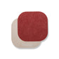 Two rounded edge square washable paper coasters lie one atop one another. The one in the foreground is red and the one at rear is beige.