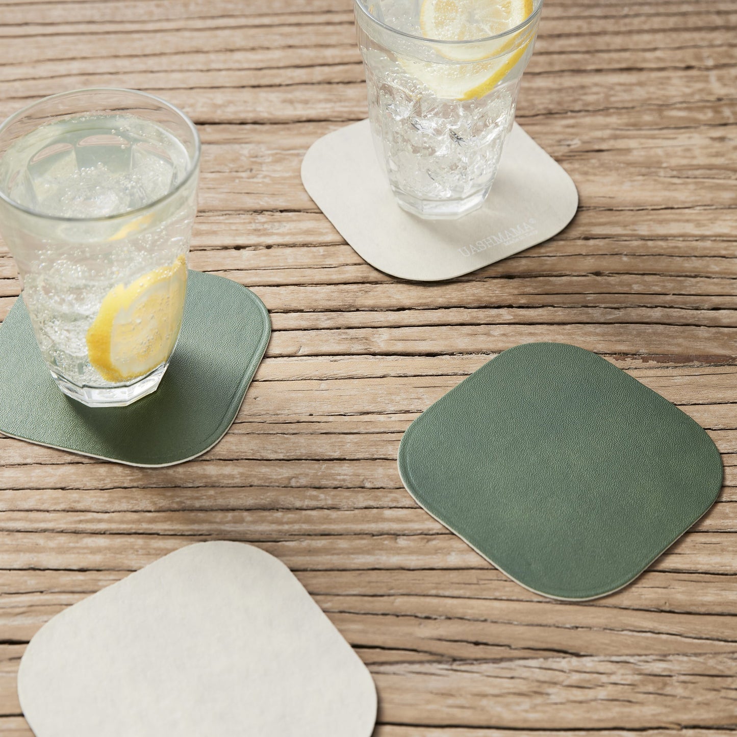 Two water glasses sit atop washable paper coasters, one in white and another in green. Another white and another green washable paper coaster sit in the foreground, atop a wooden table.