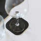 A wine glass sits on a white table, atop a black washable paper square coaster with rounded edges.