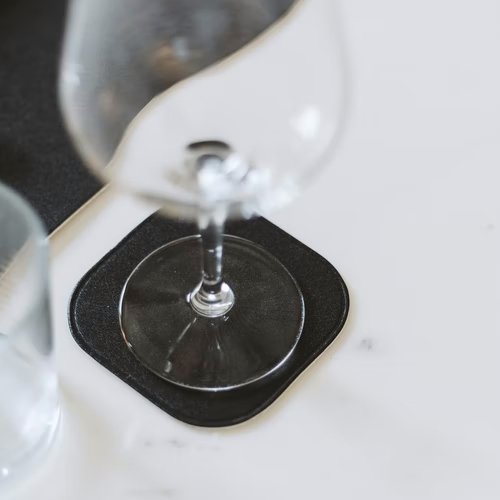 A wine glass sits on a white table, atop a black washable paper square coaster with rounded edges.