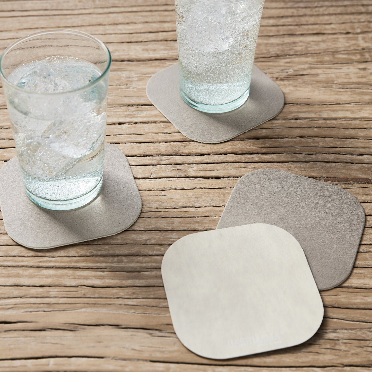 A wooden table is shown with two water glasses sitting atop grey washable paper coasters with rounded edges. At right a beige coaster overlaps another grey washable paper coaster.