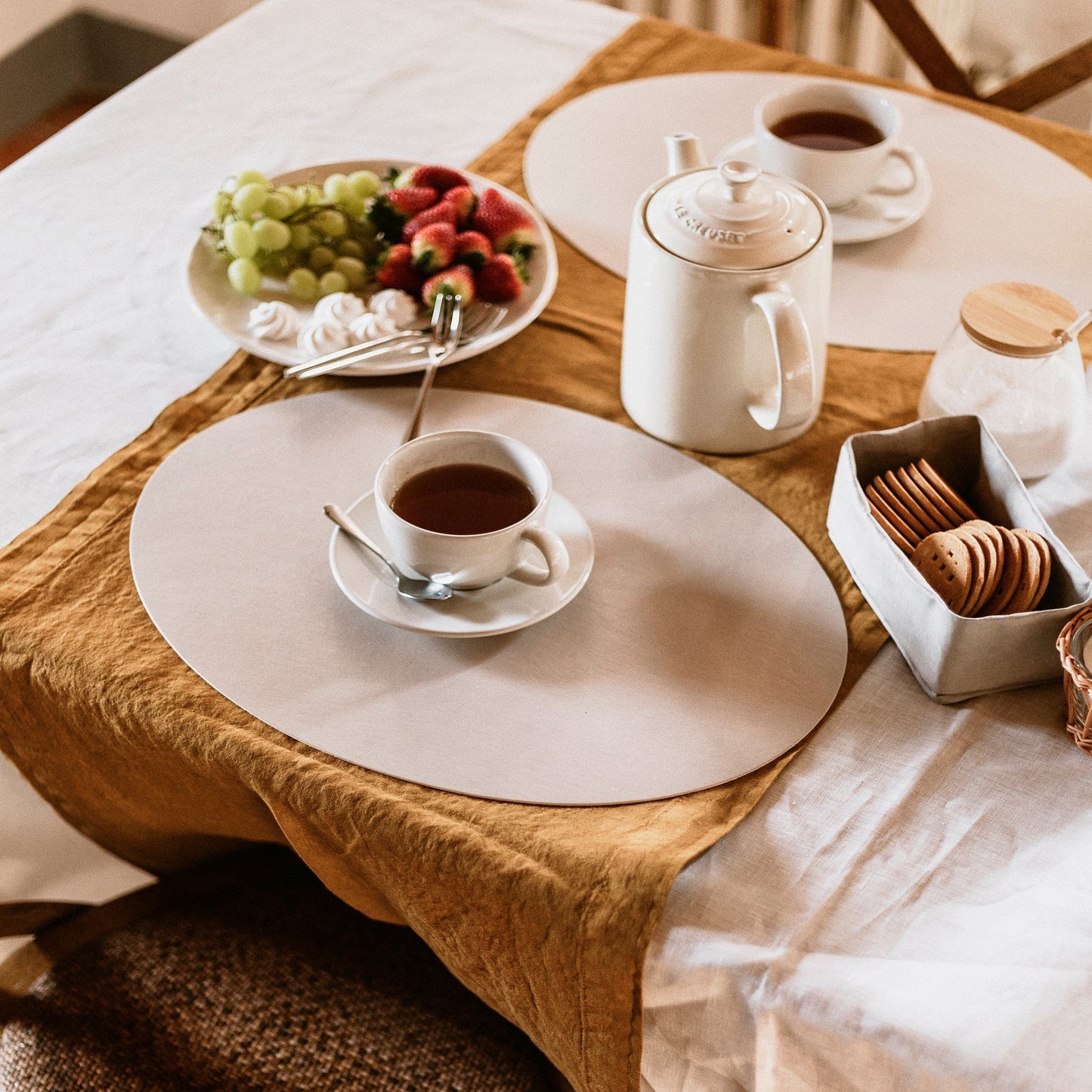 A beige oval washable paper placemat is shown atop a linen tablecloth-clad table. Atop it sits a cup of coffee, at left is a bowl of fruit, and at right is a washable paper basket holding biscuits.