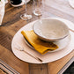 A beige washable paper oval placemat sits atop a wooden table, under a cream ceramic plate and bowl, with a mustard linen napkin and glasses in the background. 