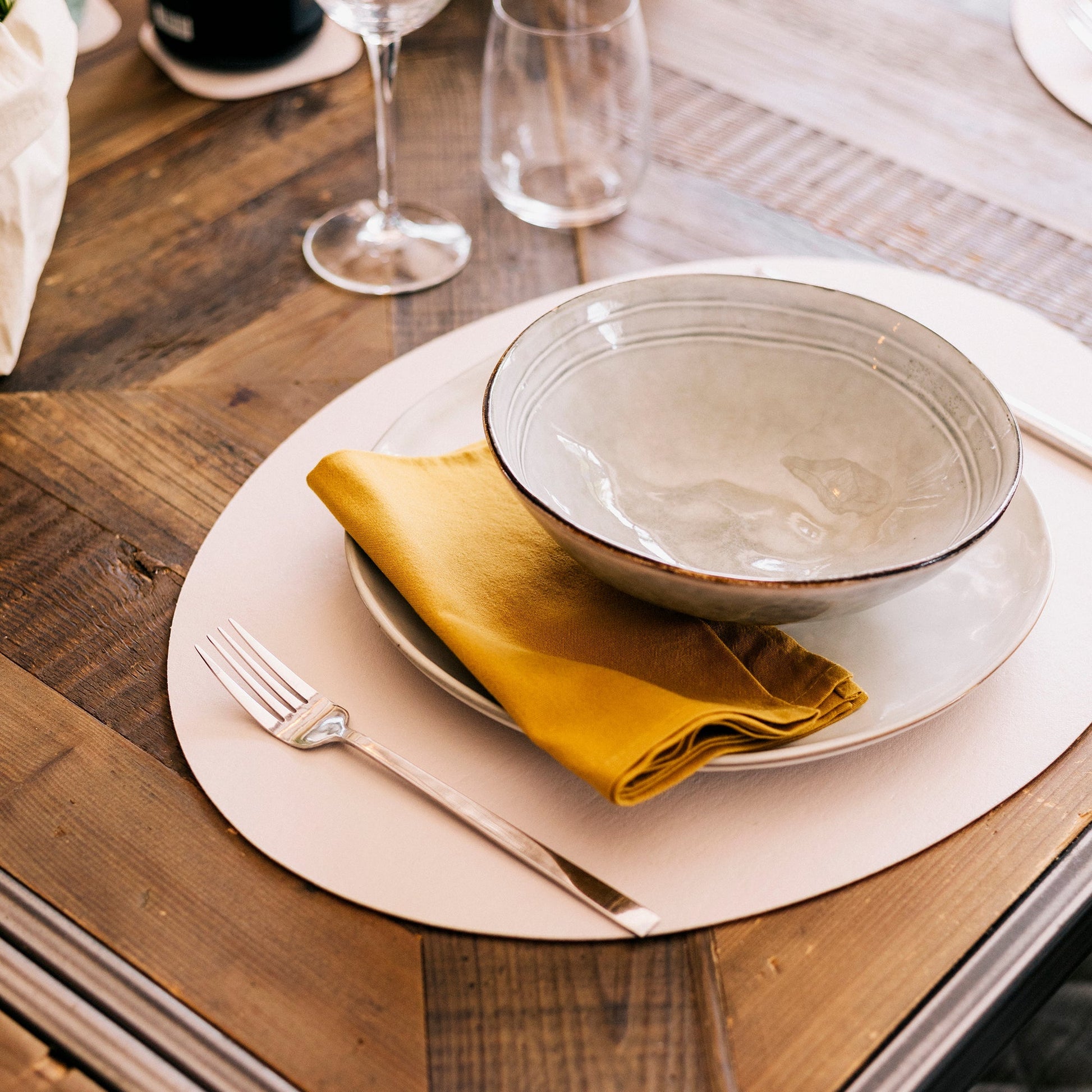 A beige washable paper oval placemat sits atop a wooden table, under a cream ceramic plate and bowl, with a mustard linen napkin and glasses in the background. 