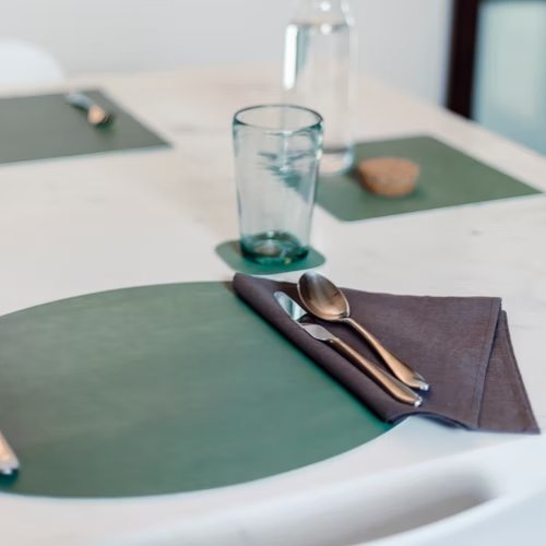 A green oval washable paper placemat is shown on a table setting, with a washable paper coaster in the same colour. A linen napkin sits atop it at right under cutlery.