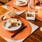 An orange oval washable paper placemat sits atop a beige rectangular washable paper placemat on a wooden table. The table is set for breakfast, showing toast, cereal and orange juice. Walnuts sit in a washable paper square basket in the background.