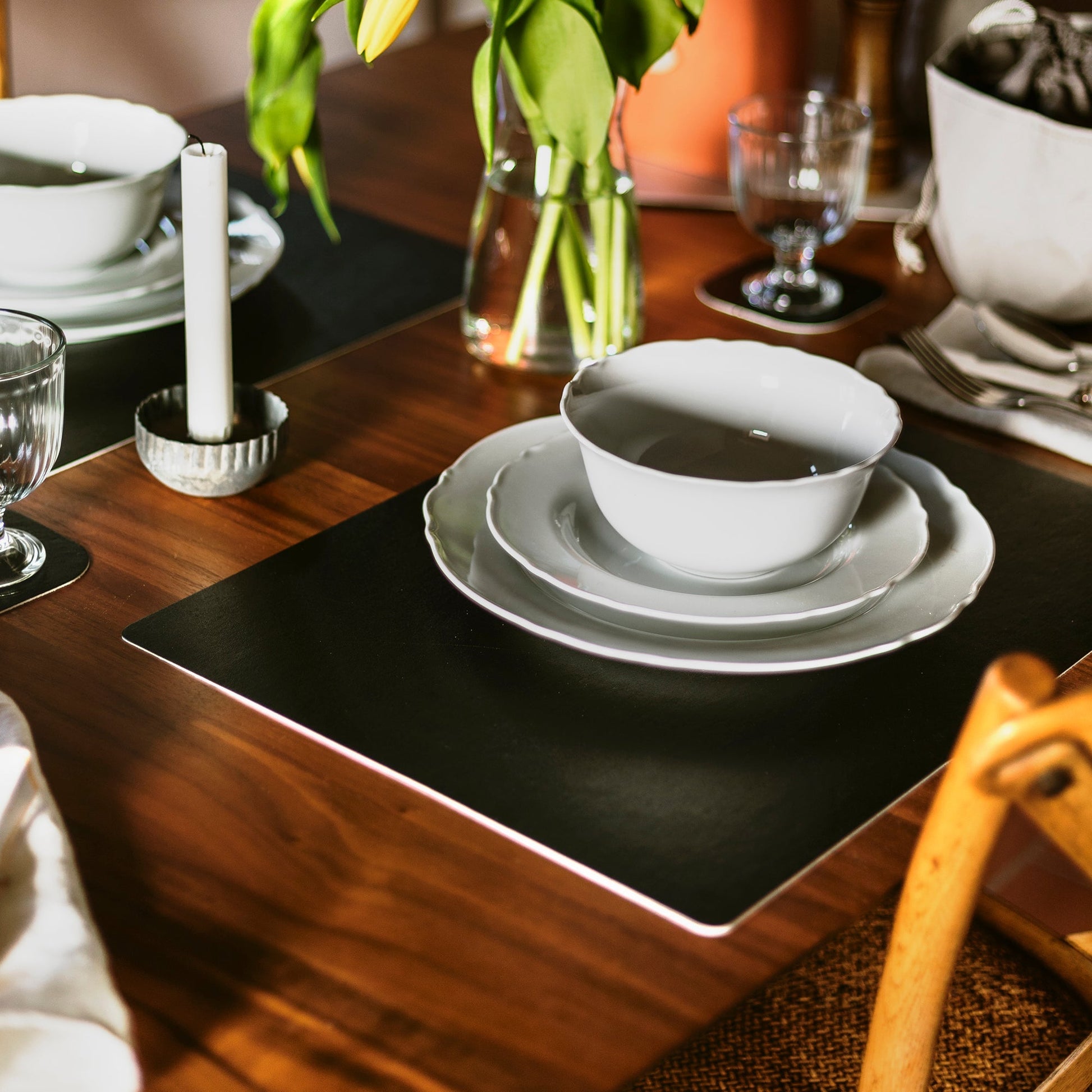 A black washable paper placemat is shown on a wooden table setting, underneath a stack of white ceramic plates and a bowl.