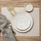 A beige washable paper placemat is shown on a wooden table. On top of it are two white ceramic plates, a ceramic dish, a water glass, a grey linen napkin and a fork and spoon.