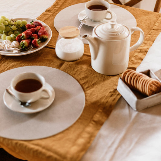 Two round beige washable paper placemats are shown on a breakfast table setting, with ceramic cups of coffee, a coffee pot, a bowl of fruit and a washable paper small square basket containing biscuits.