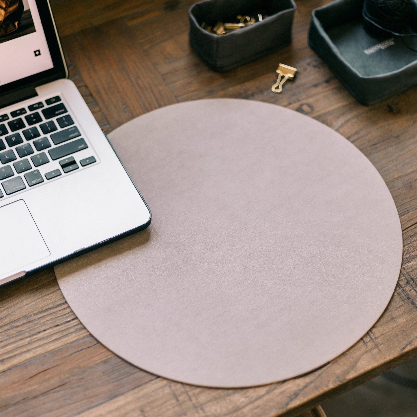 A beige round washable paper placemat is shown on a desk setting, under a laptop. At right are black washable paper small baskets containing stationery clips.