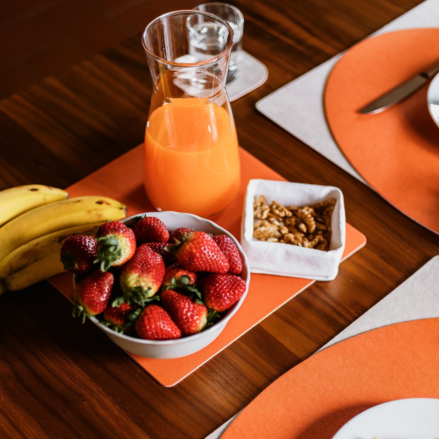 Three orange washable paper placemats sit on a wooden table in a place setting. The hero of the image is a square orange washable paper placemat containing bananas, a carafe of orange juice, a bowl of strawberries and a washable paper small tray containing nuts.