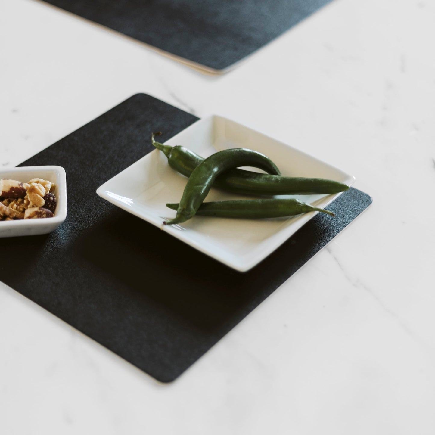 A black square washable paper placemat sits atop a white table. On top of it is a small ceramic plate with green peppers, and a small ceramic plate containing nuts.