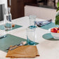 A white table is shown laid with four green washable paper placemats - one is square, one rectangular, one round and one oval.