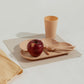 A beige square washable paper placemat sits on a white table. On top of it is a child's plate, cup, and cutlery in matching melamine peach colour, with an apple on the plate.
