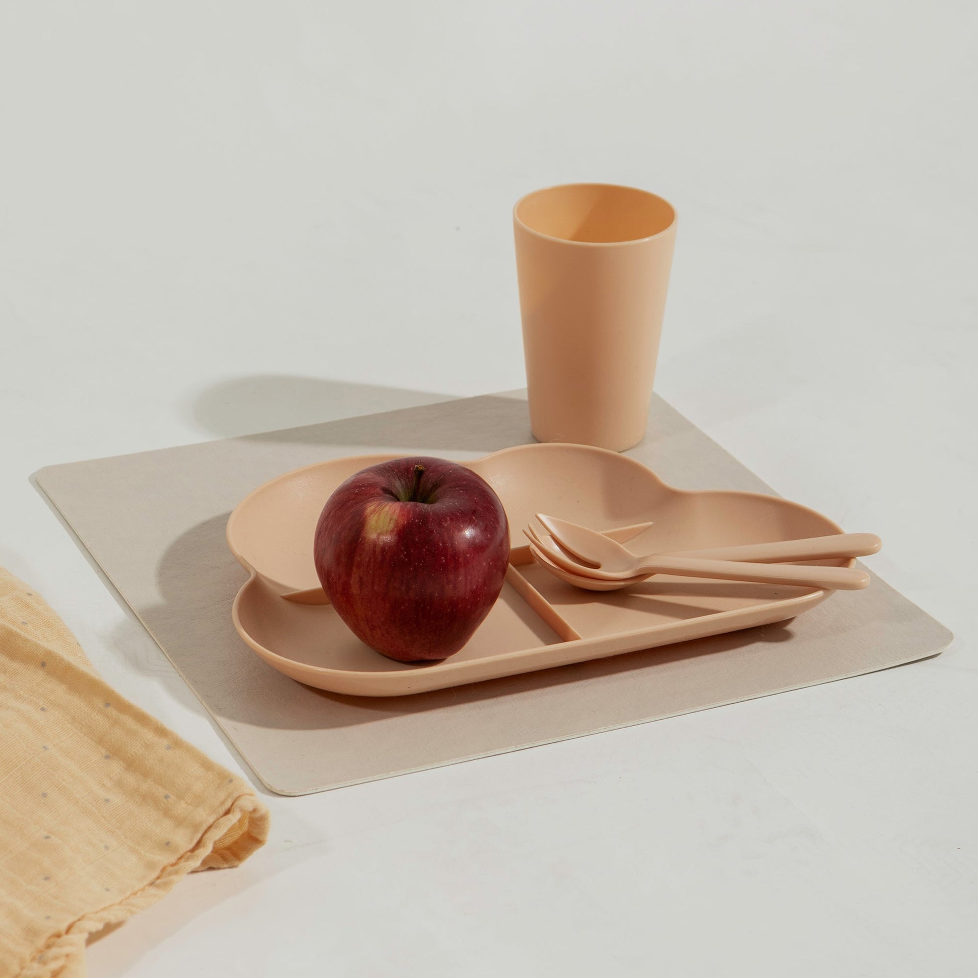 A beige square washable paper placemat sits on a white table. On top of it is a child's plate, cup, and cutlery in matching melamine peach colour, with an apple on the plate.