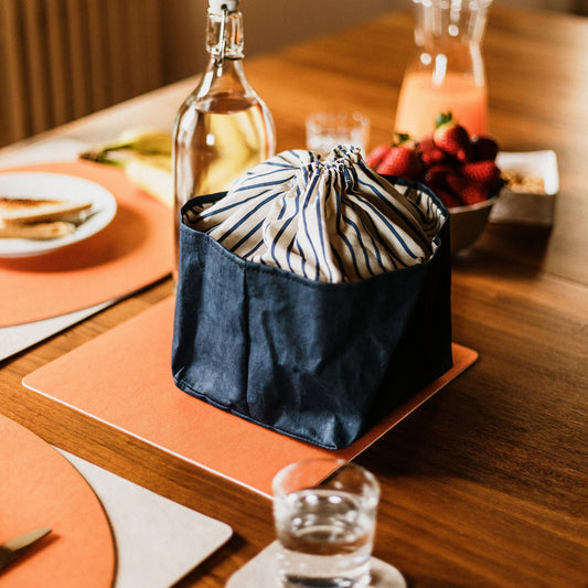 A square washable paper orange placemat sits on a dining table, underneath a washable paper bread basket with a drawstring top.