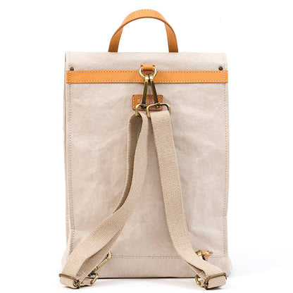 A cream washable paper backpack is shown from the back angle. It features a tan strip across the back, a tan top handle, and cream cotton canvas straps with gold metallic hardware.