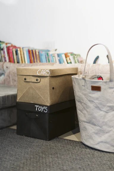 Two washable paper boxes are shown in a home setting, one labelled "cars" and the other labelled "toys." They are the same size, one is in beige and one is in black. At right sits a white washable paper tote bag. 