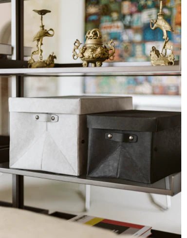 A set of two washable paper lidded boxes are shown on a shelf in a home setting. The one at left is larger and grey and the one at right is smaller and black.
