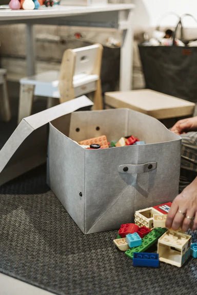 A grey washable paper basket is shown in a home setting. The lid is off, and a woman's hands are shown filling it with lego.