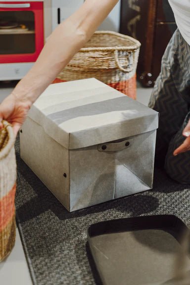A washable paper grey lidded box is shown in a home setting. A woman is shown leaning over it. 