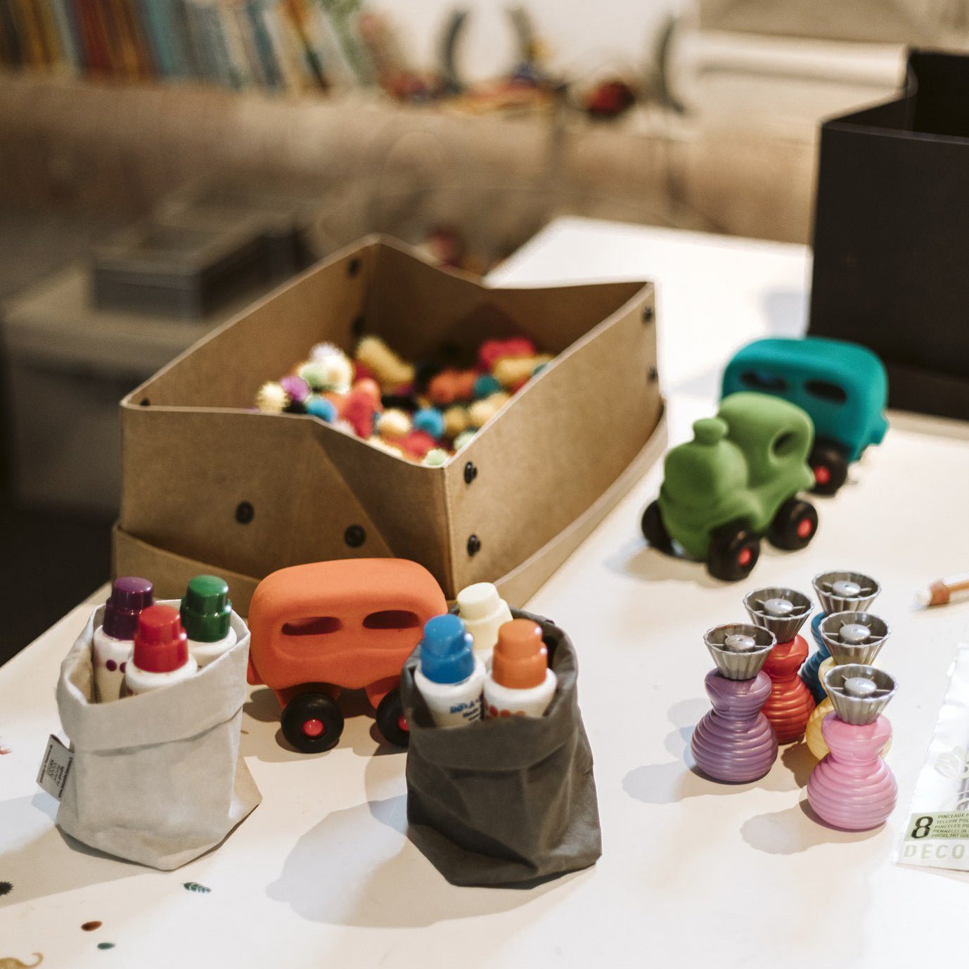 A tan washable paper box is shown sitting inside its lid, filled with toys. Two washable paper small bags are shown holding children's paints in the foreground.