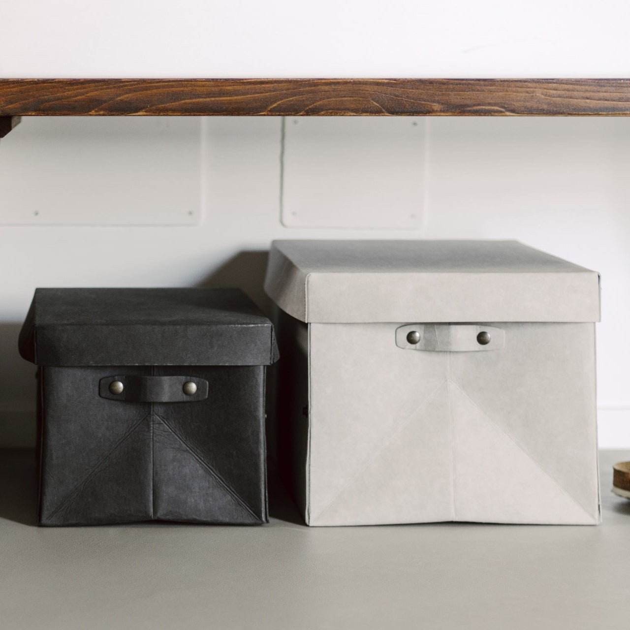 Two washable paper lidded boxes are shown in a home setting. The one at left is smaller and black and the one at right is grey.