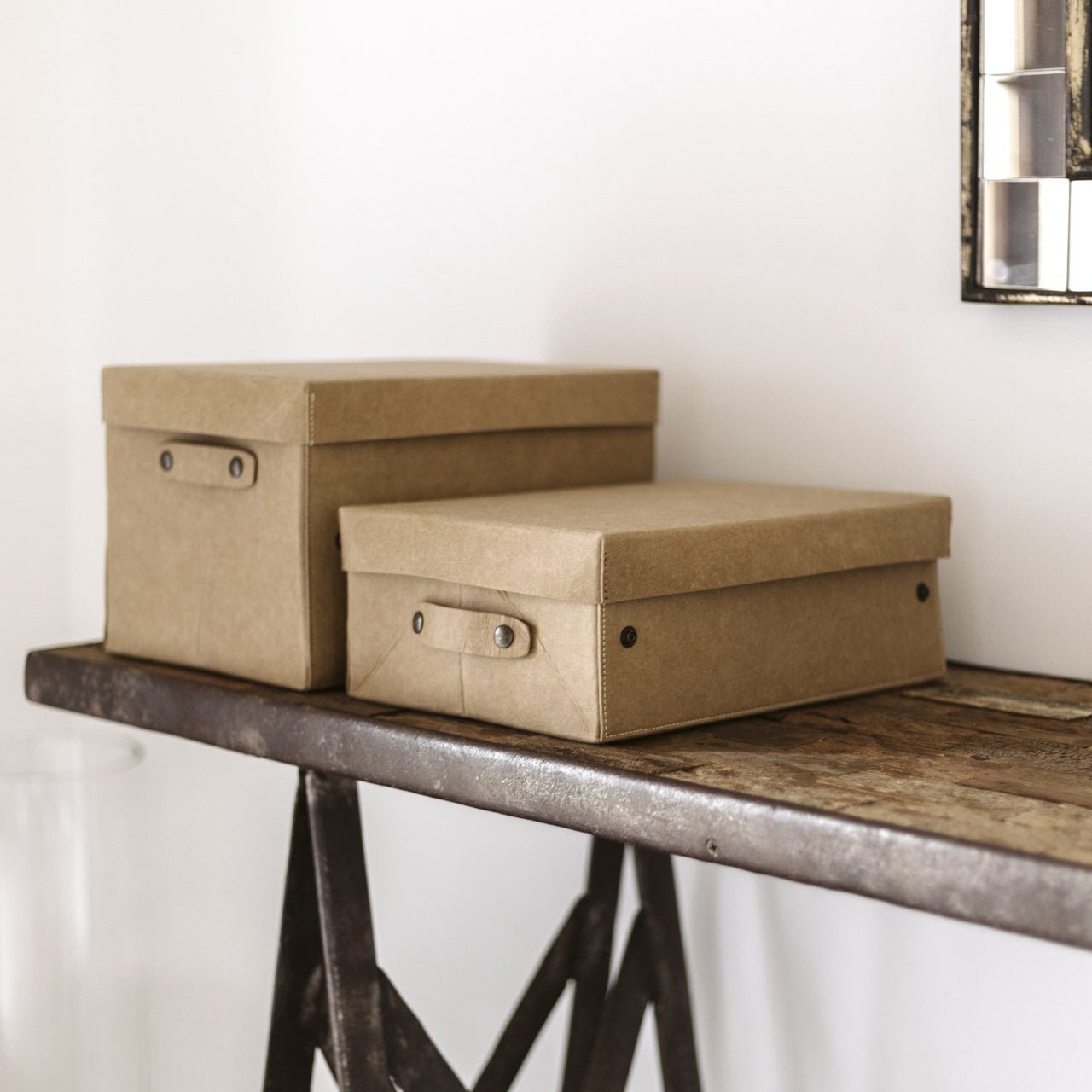 Two tan washable paper lidded boxes are shown on a metal and wood console table in a home setting, under a mirror.