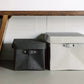 Two washable paper lidded boxes are shown sitting on the floor under a bench. The one at left is smaller and black and the one at right is grey.
