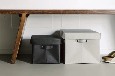 Two washable paper lidded boxes are shown sitting on the floor under a bench. The one at left is smaller and black and the one at right is grey.
