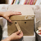 A woman's hands are shown attaching the metal stud closure handle to a tan washable paper box labelled "cars." It sits atop a grey, larger washable paper lidded box.