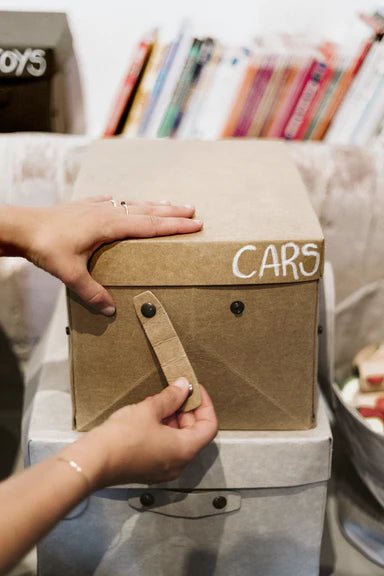 A woman's hands are shown attaching the metal stud closure handle to a tan washable paper box labelled "cars." It sits atop a grey, larger washable paper lidded box.