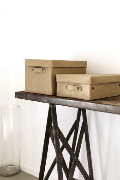 Two tan washable paper lidded boxes are shown on a metal and wood console table in a home setting.