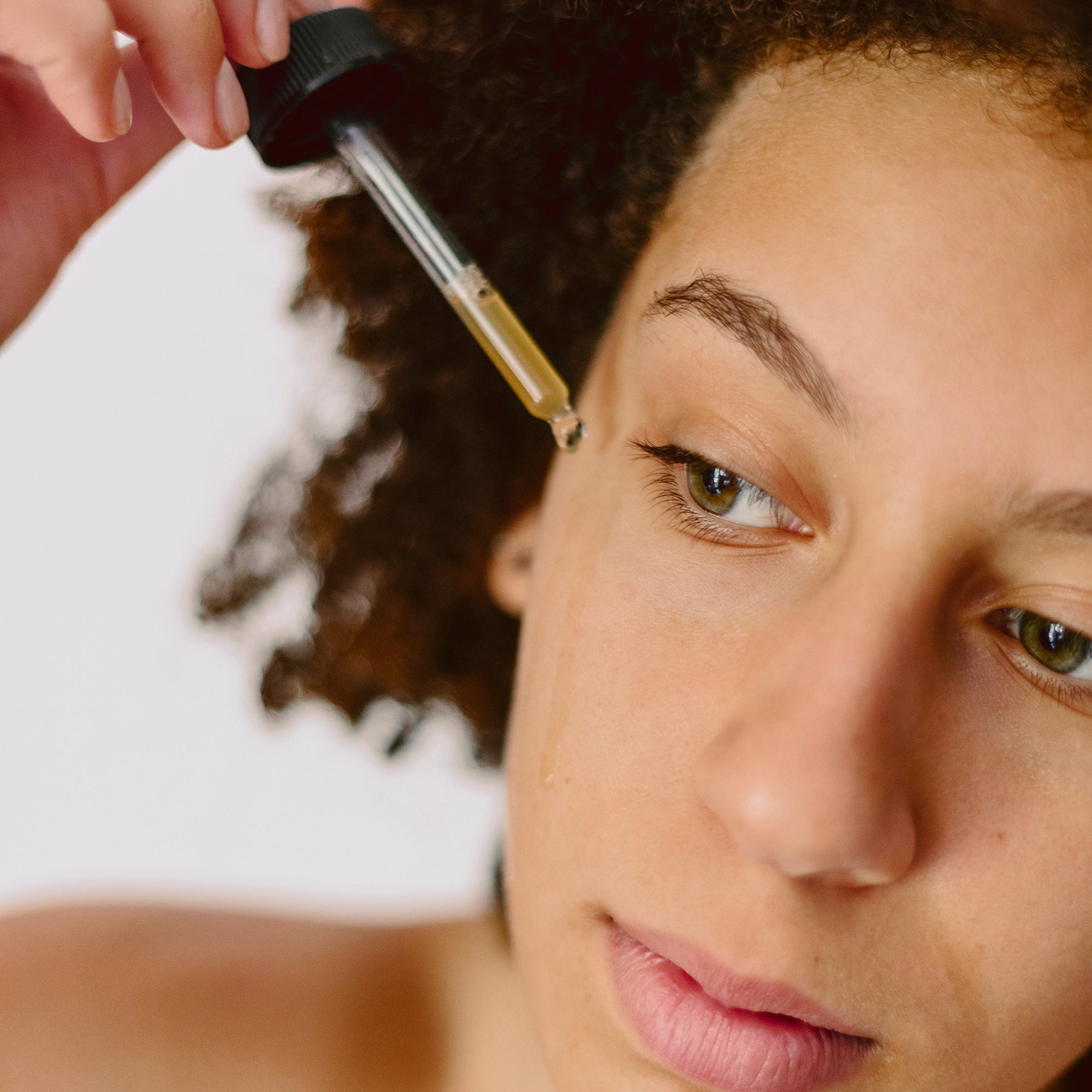 A woman is shown applying a serum to the eye area using a glass dropper.