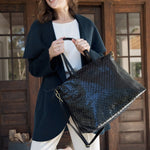 A woman stands outside a house wearing a navy cardigan, white t-shirt and chinos. She carries a large black woven washable paper tote by its two top handles.