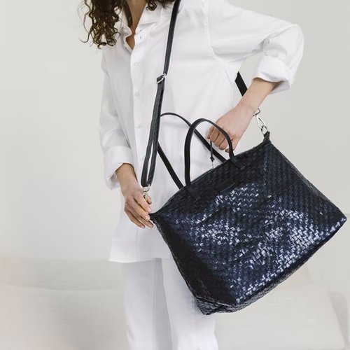 A woman dressed in white wears a large shiny navy metallic woven washable paper tote across one shoulder, by way of a dark canvas strap. It features two top handles.