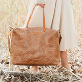 A woman in cream is shown walking through a field at a close up angle. She carries a large tan woven washable paper tote by its top handles.