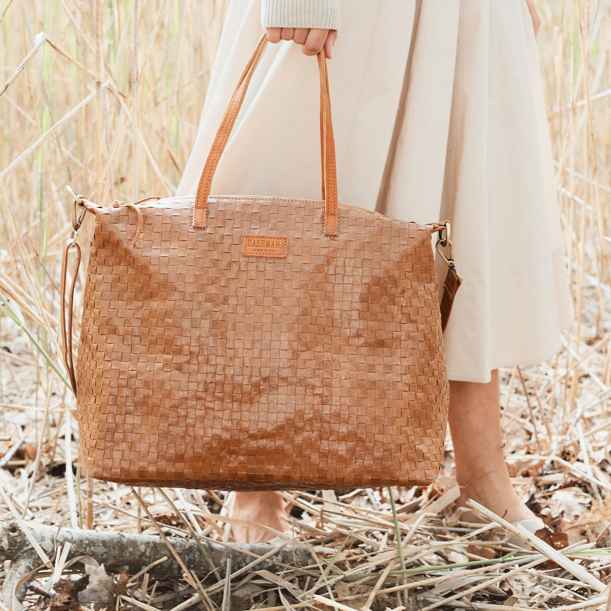 A woman in cream is shown walking through a field at a close up angle. She carries a large tan woven washable paper tote by its top handles.