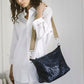 A woman dressed in white carries a woven washable paper handbag over one shoulder. It is metallic navy, with a beige canvas strap and silver hardware.