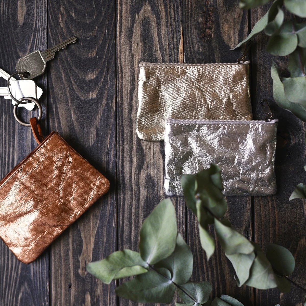 Three washable paper small pouches sit on a wooden table top. The one at left shows three keys attached to its inner keyring. They are from left to right metallic rose gold, metallic gold, and metallic silver.