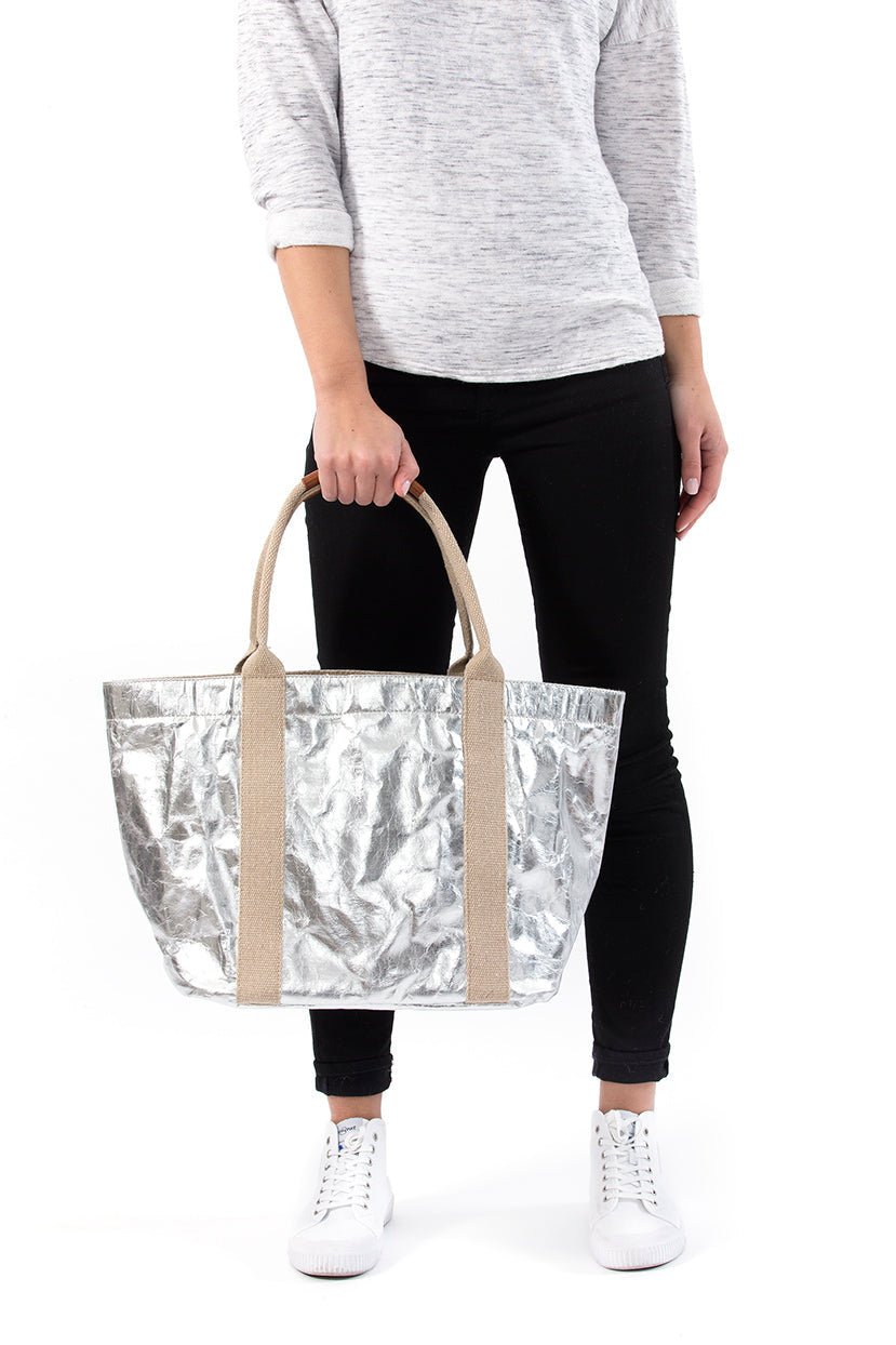A woman in casual clothing is shown holding a silver metallic washable paper tote bag in front of her with two beige cotton canvas top handles.