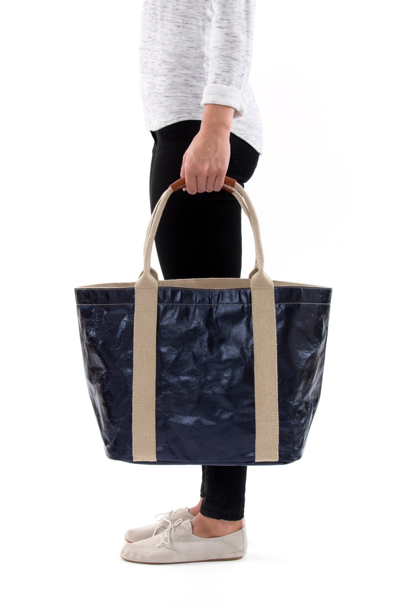 A woman in casual clothing is shown holding a navy metallic washable paper tote bag with two beige cotton canvas top handles.
