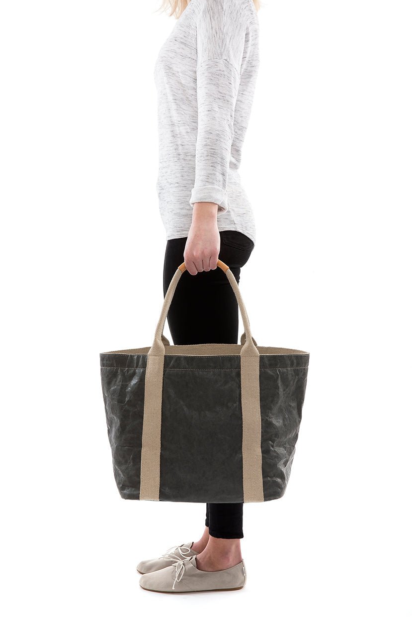 A woman in casual clothing is shown holding a dark grey washable paper tote bag with two beige cotton canvas top handles.