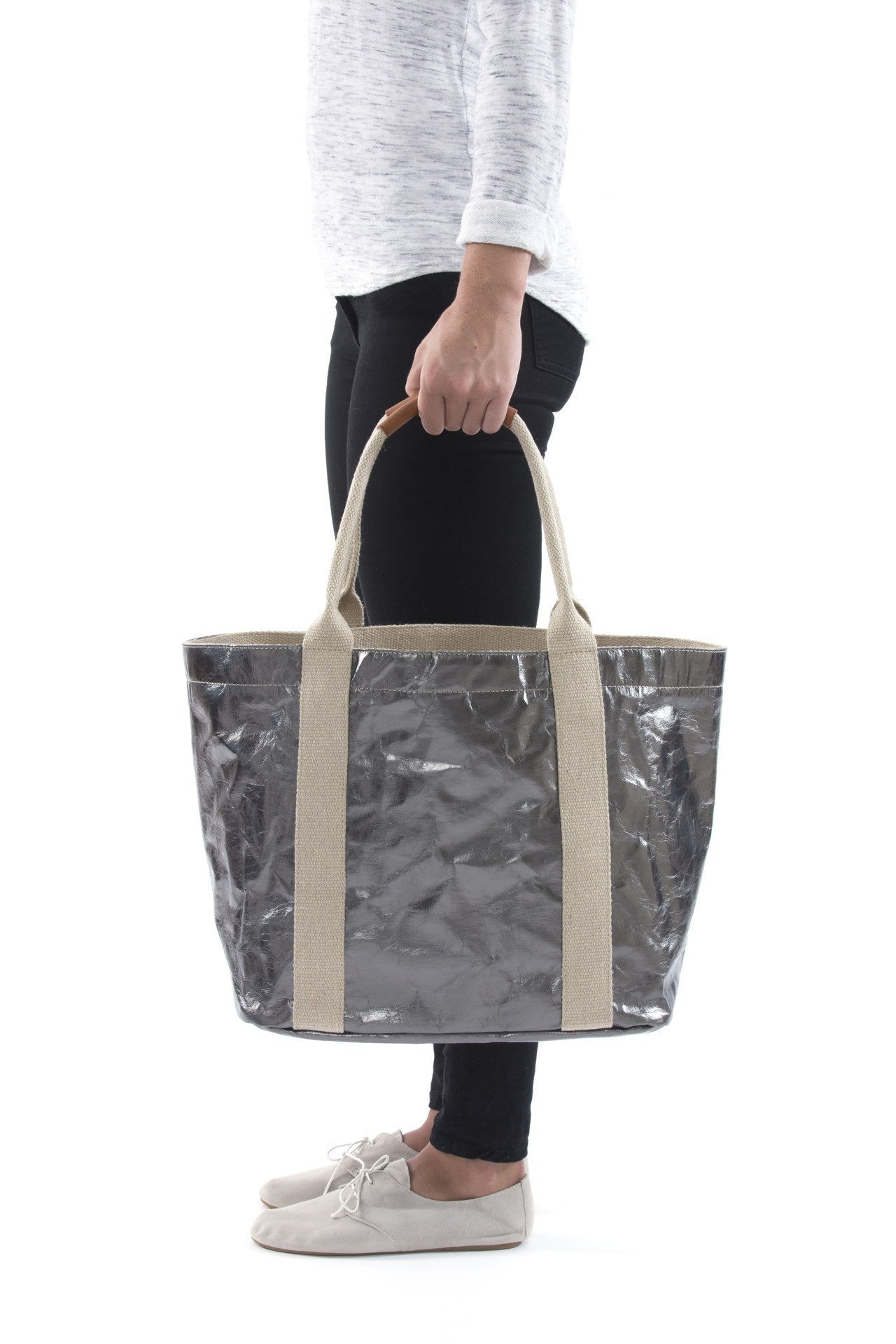 A woman in casual clothing is shown holding a metallic dark silver washable paper tote bag with two beige cotton canvas top handles.
