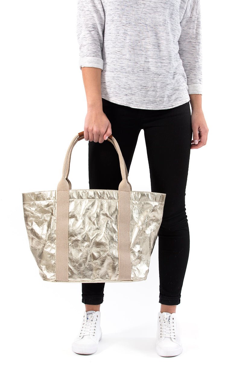 A woman in casual clothing is shown holding a gold metallic washable paper tote bag in front of her with two beige cotton canvas top handles.