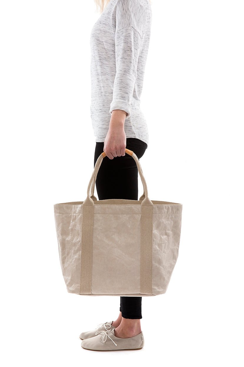 A woman in casual clothing is shown holding a cream washable paper tote bag with two beige cotton canvas top handles.